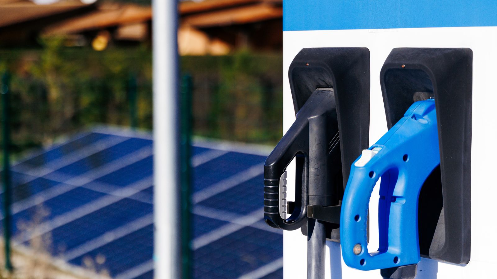 Connectors of an electric vehicle charger with solar panels in the background.